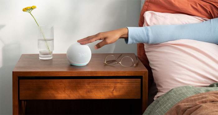 Amazon Echo Dot Gen 5 on nightstand with flower vase, glasses and hand reaching toward the Echo Dot.