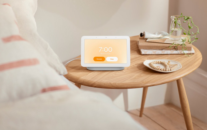 Google Nest Hub 2nd gen on wooden table next to a bed. On the table are books, a dish with jewelry and a vase with a plant.