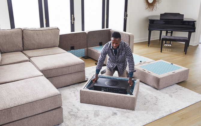 Man placing subwoofer into the bottom of a Sactional seat. In the background the other pieces of the Sactional with three additional bases. To the right is a piano. 