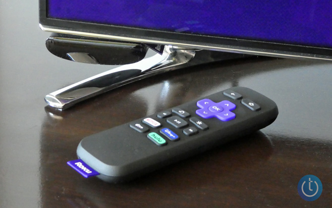 Roku Express mounted on the bottom of a TV with the remote on a wooden console.