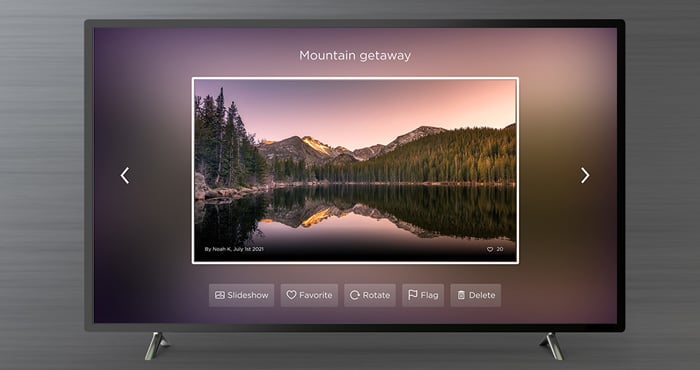 Roku Photo Streams feature on TV sitting on a gray background. You see a mountain landscape and menu items below: Slideshow, Favorite, Rotate, Flag, Delete.