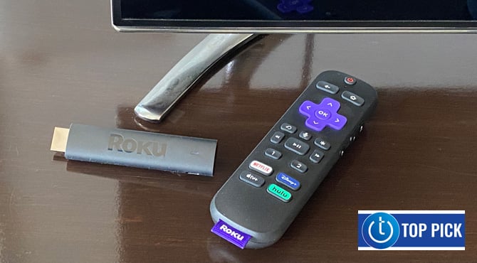 Roku Streaming Stick 4K+ with remote control on wooden table with TV. The Techlicious Top Pick Award logo is in the lower right corner.
