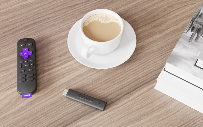 Roku Voice Remote Pro on the left, with a coffee cup in the middle, the Roku Streaming Stick 4k Plus below the coffee cup and a black-and-white print on the right side. Everything is on a wooden table. 