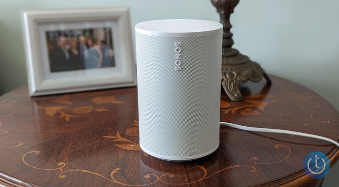 Sonos Era 100 on a table with a picture frame and lamp for scale.