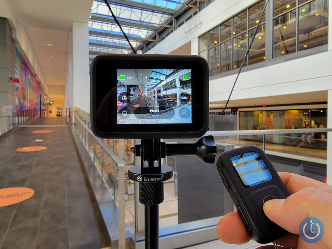 GoPro Hero10 Black on selfie stick in the hallway of a shopping mall with glass ceilings. Also in the photo is a hand holding The Remote. 