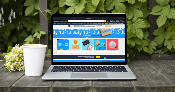 Macbook Air on wooden surface with coffee cup and a background of plants. The computer screen has a screenshot of Amazon website showing Amazon Prime Day. 