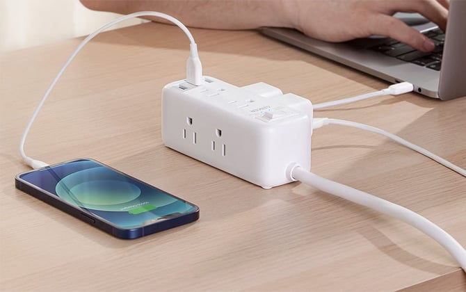 Anker PowerExtend USB Surge Protector shown on a wood desk with a phone plugged into a USB port, two power bricks on the back, one of which is plugged in a partially visible laptop. You see an arm and hand on the laptop keyboard. 