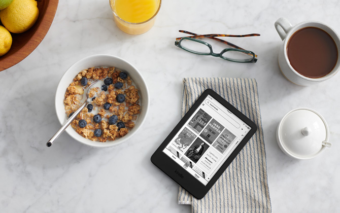 Kindle 11th generation shown on a dish towel with a bowl of cereal, sugar bowl, juice glass, coffee, bowl or fruit and glasses for size comparison.