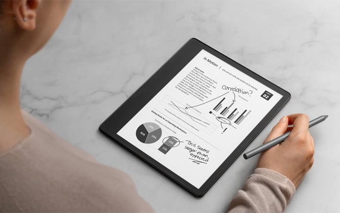 Kindle Scribe shown with a textbook page and hand-written notes. A person is seated next to it holding the pen.