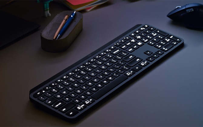 Logitech MX Keys in dark room on desk with backlight on. To the left on the desk is a pencil holder and in the top right is a mouse.