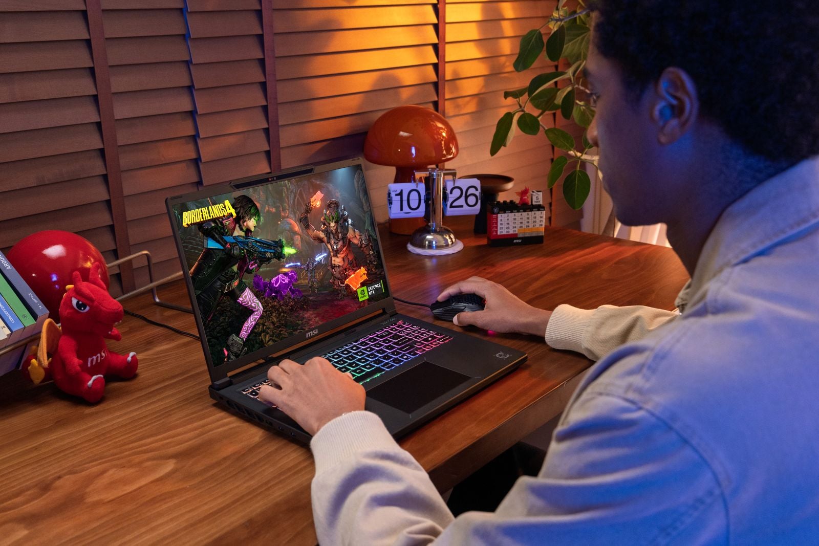 A person plays Borderlands 4 on an MSI Crosshair A16 HX laptop at a wooden desk, one hand on the RGB-lit keyboard and the other on a gaming mouse.