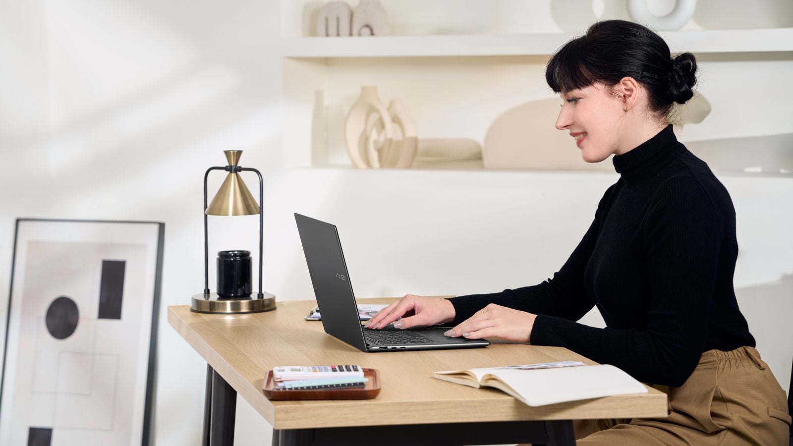 Woman in a black turtleneck typing on an MSI Prestige 16 AI+ laptop at a wooden desk in a bright, minimalist home office