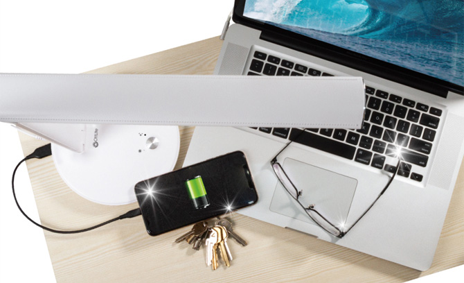 OttLite Emerge desk lamp on wooden desk shown from the top down. The light is over a laptop keyboard that has a phone, keys and glasses on it.  The phone is plugged into the USB-A port at the back of the light.