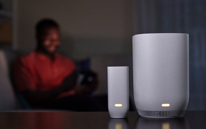 Comcast xFinity Storm-Ready WiFi device shown in a dark room with a man reading in the backgraound. On the left is the slimmer battery pack, on the right is the Storm-Ready.