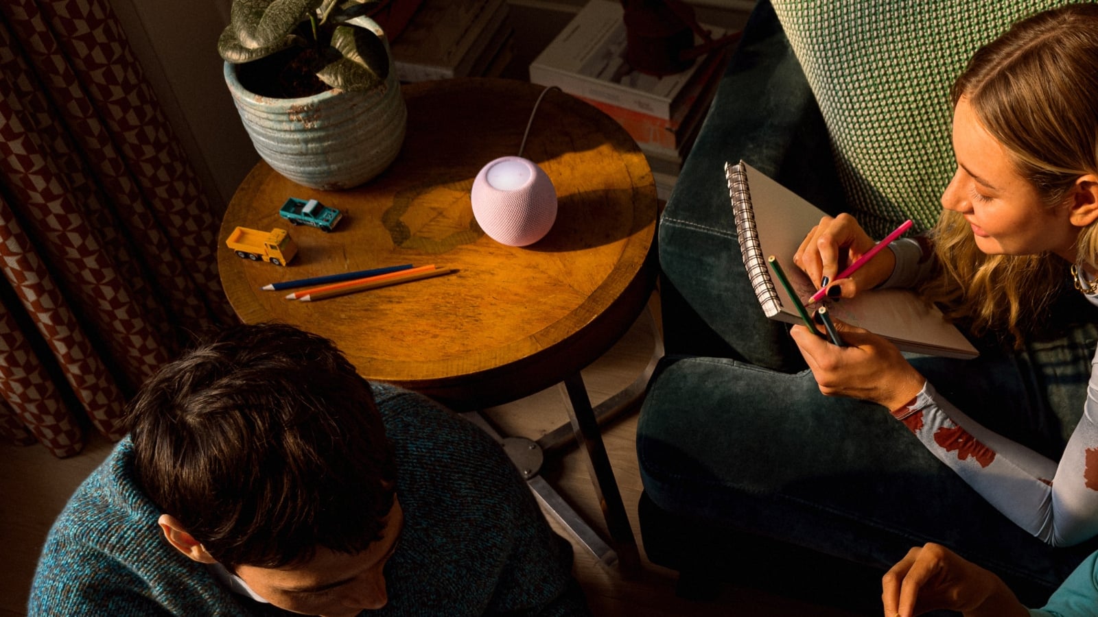ink Apple HomePod mini 2 on a wooden side table beside a woman drawing in a sketchbook, with colored pencils and toy cars nearby.