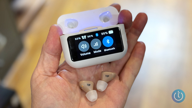 The Audien Atom X are held in a hand with the case. The case shows volume, mode, and bluetooth buttons.