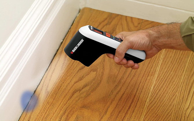 Black & Decker TLD100 Thermal Leak Detector held in hand pointed at baseboard. A blue dot is shown on the baseboard to show where the detector is measuring.