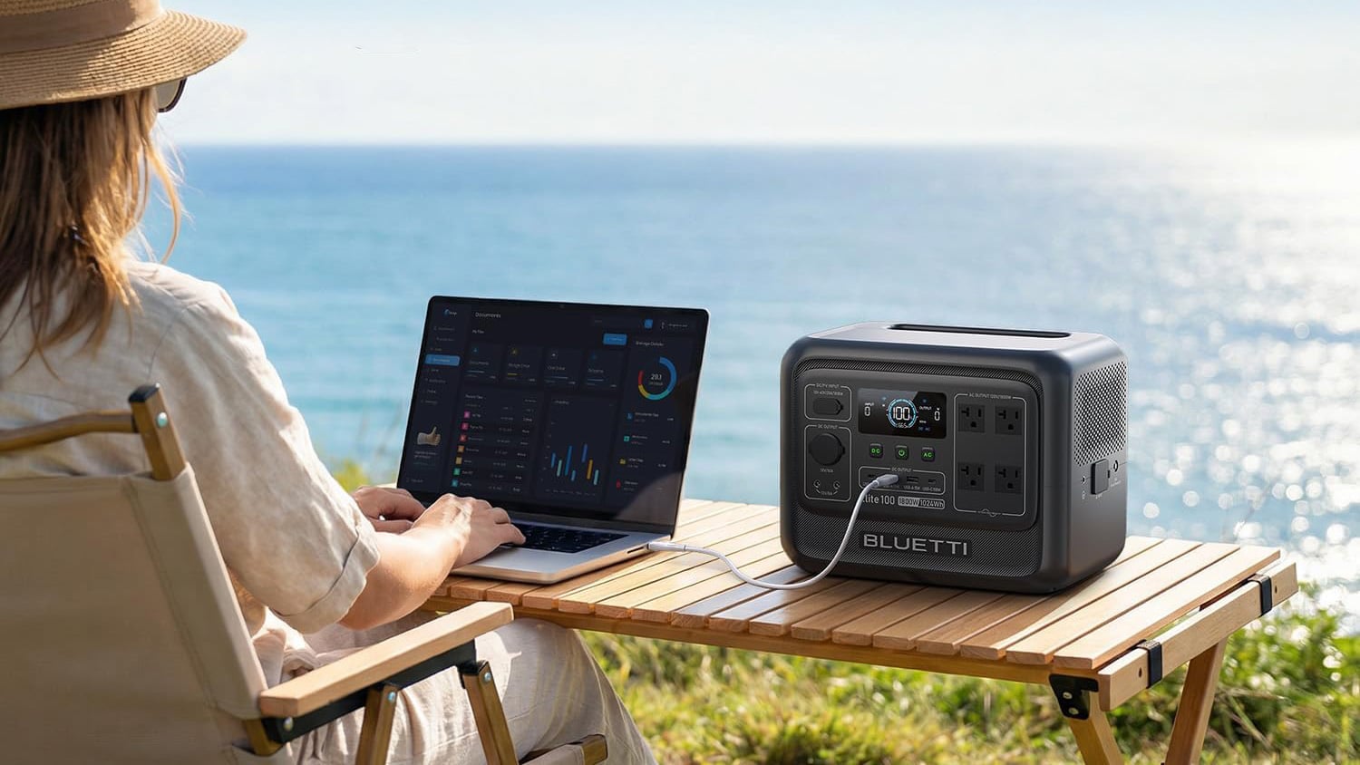 BLUETTI Elite 100 V2 portable power station connected to a laptop on an outdoor table overlooking the ocean.