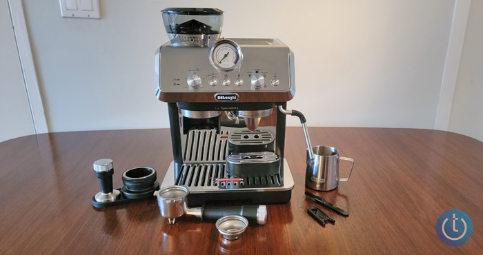 DeLonghi La Specialista Arte on wood table with all of the accessories: dosing funnel, tamper, and a mat to hold the filter while you're tamping, milk frother, single-shot basket, double-shot basket, portafilter, cleaning brush, and cleaning tool for milk frother. 