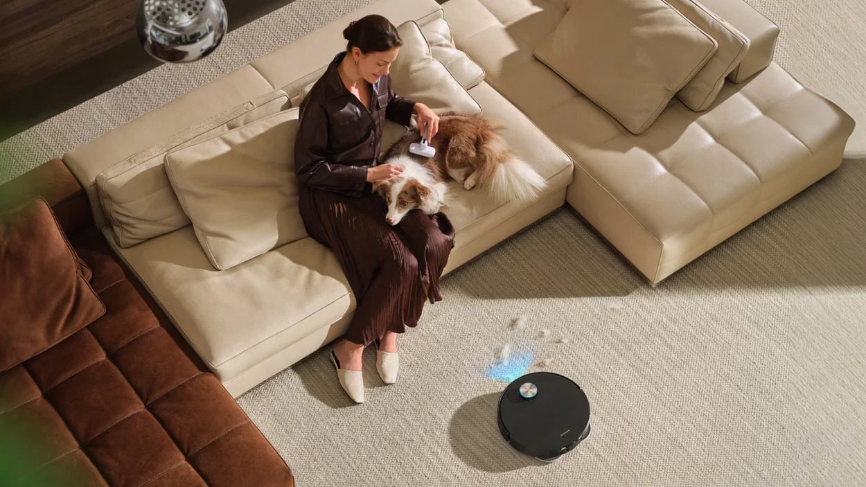 Dreame X60 Ultra robot vacuum cleaning near a sofa where a woman sits with her dog.
