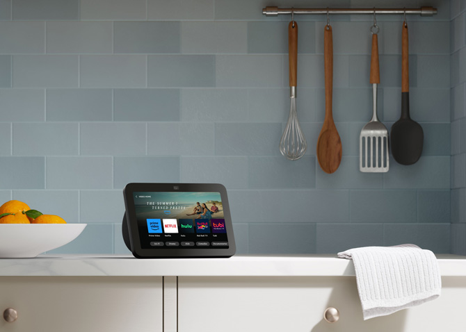 Amazon Echo Show 8 Gen 3 shown on a kitchen counter with hanging kitchen utensils and a bowl of oranges for scale.