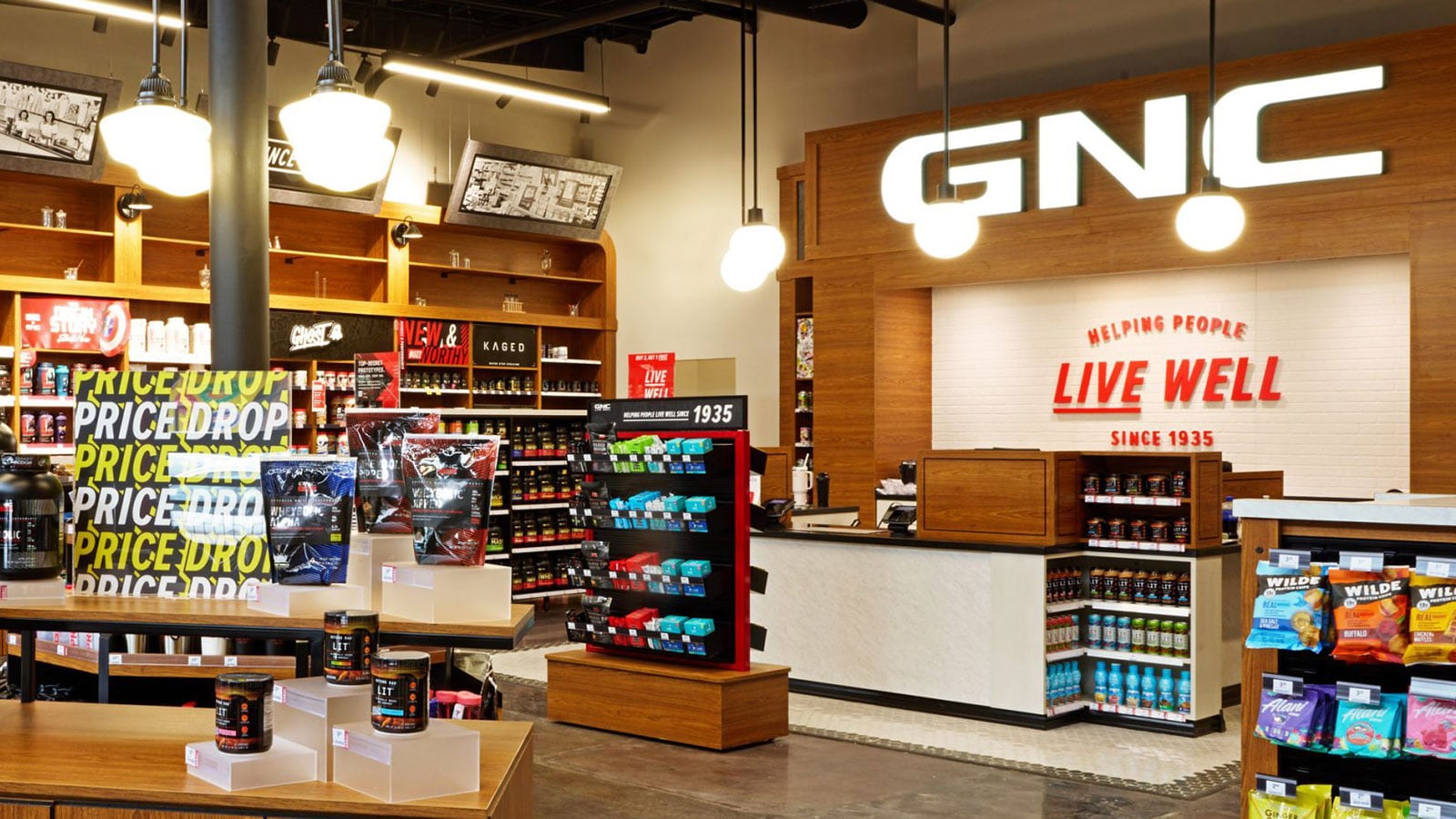 Interior of a GNC store with shelves and displays of vitamins, protein powders, and supplements, beneath a large illuminated GNC logo.