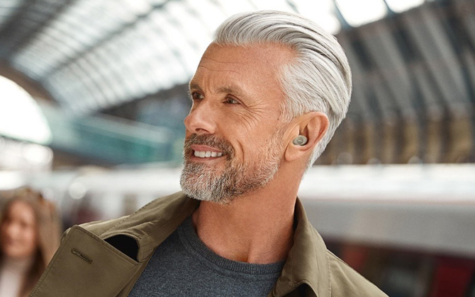Jabra Enhance Plus Self-Fitting OTC buds worn by man shown from the side in a subway station with people in the background.