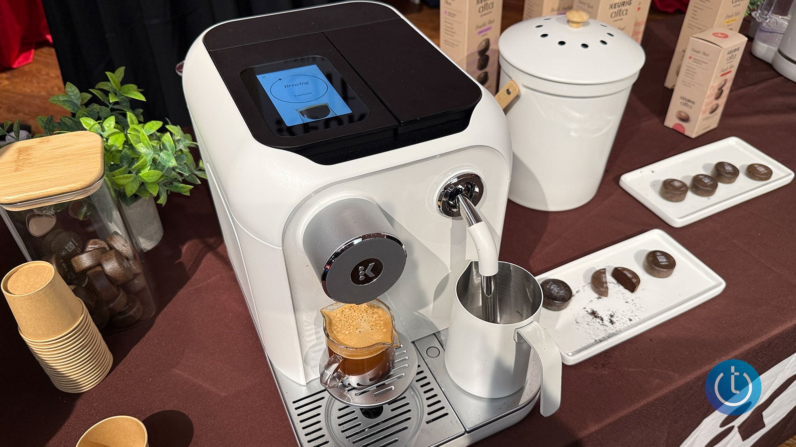 An overhead view of the Keurig Alta espresso machine with its touchscreen showing brewing in progress, alongside a milk frothing pitcher and coffee pods.