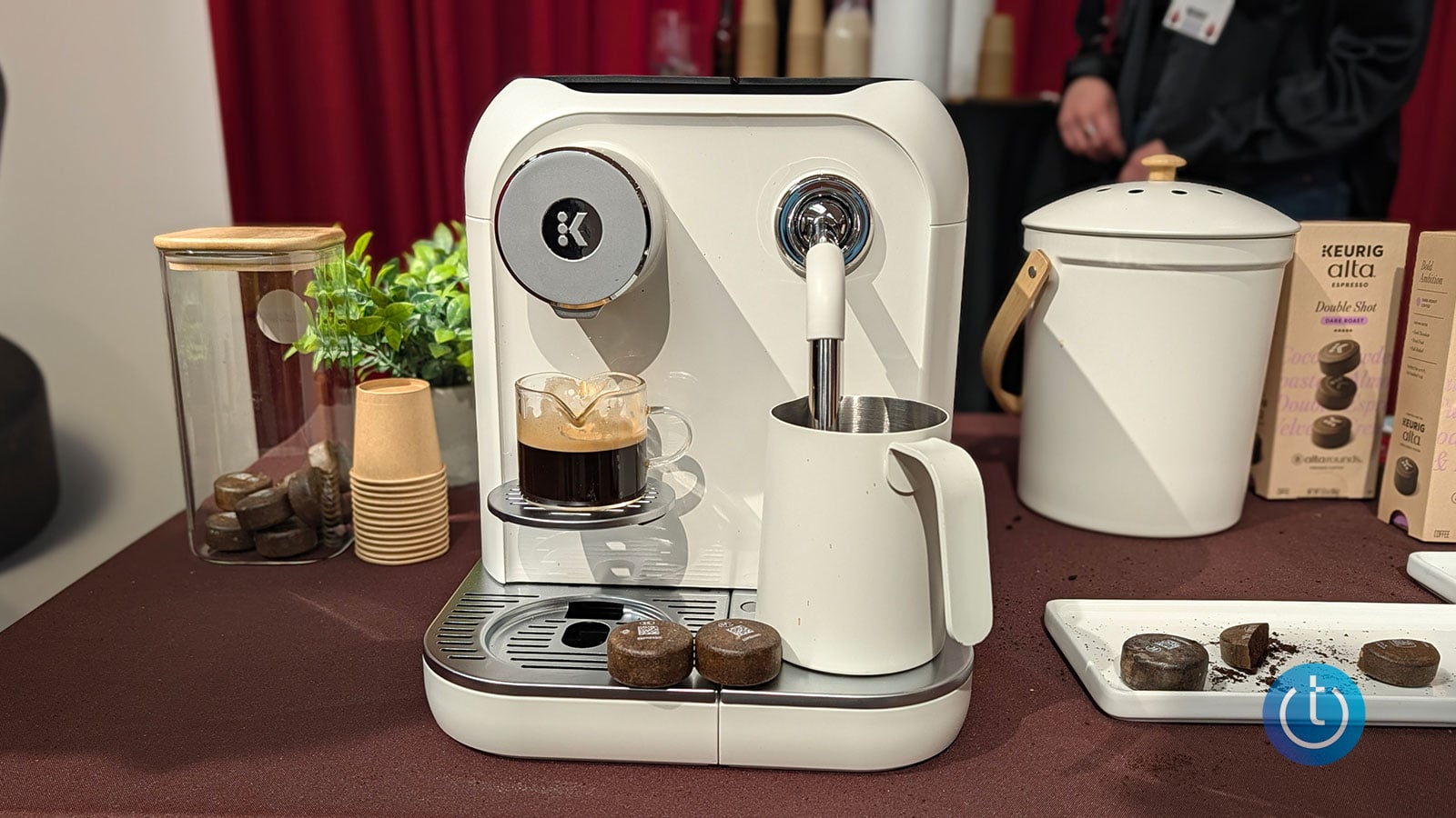 Front view of the Keurig Alta espresso machine with a freshly brewed espresso, milk frothing pitcher, and Alta coffee pods.