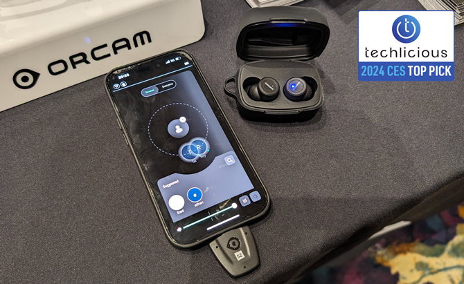 OrCam Hear OTC Hearing Aids in their case with the dongle and a phone showing two people with active audio profiles. The Techlicious 2024 CES Top Pick award logo is in the upper right corner
