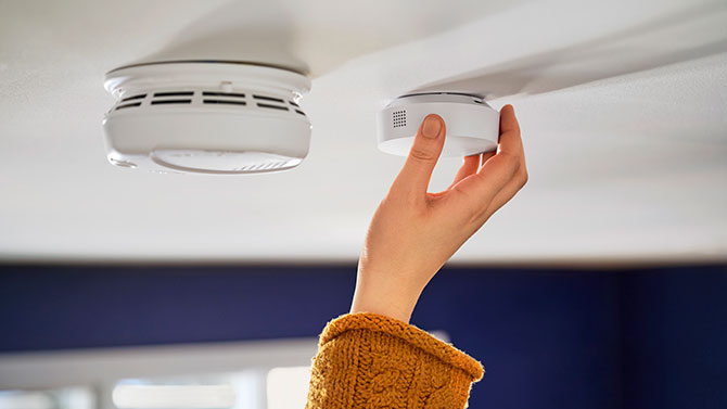 A smoke and CO detector on the left with a hand installing the SimpliSafe Listener on the right