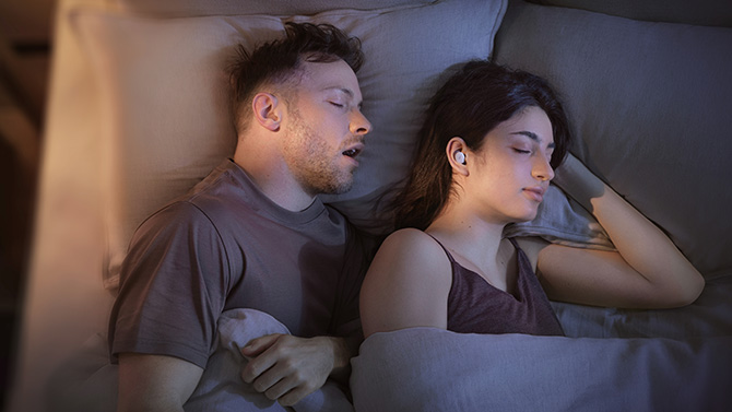 A woman wears the soundcore Sleep A30 Special in bed next to a man snoring.