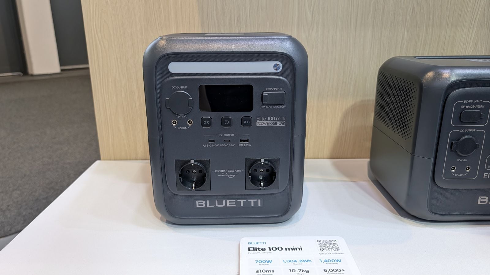 Bluetti Elite 100 mini portable power station showing its front panel with DC and AC output ports, USB-C and USB-A charging ports, and a digital display, next to a spec card listing 700W output and 1,004.8Wh capacity