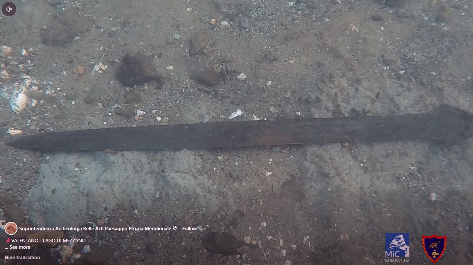 Dark bronze sword lying on the pebbly bed of Lake Mezzano in Italy, tip pointing left, surrounded by small stones and shells.