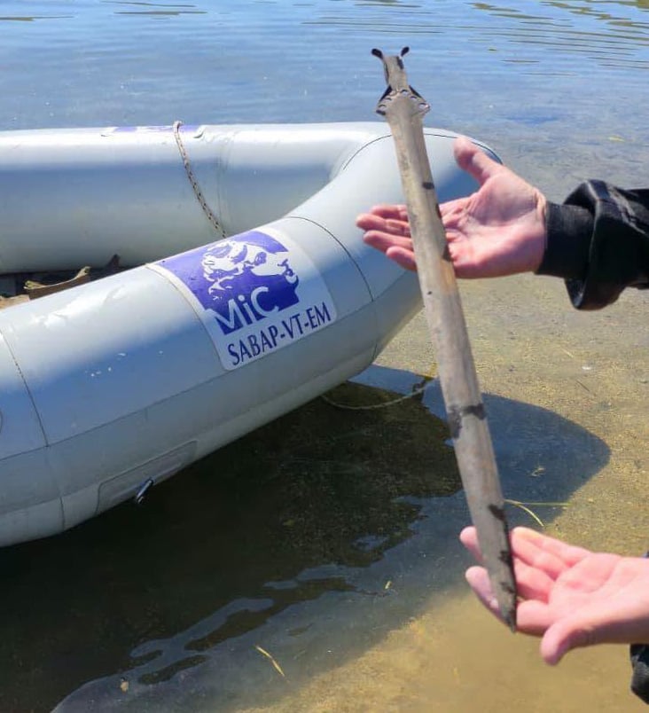 Two hands hold a dark bronze sword upright at the water's edge beside an inflatable boat marked MiC SABAP-VT-EM.