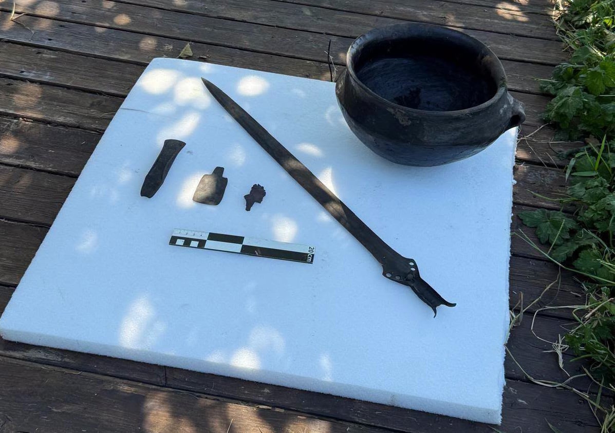Bronze sword with a swallowtail-shaped hilt end and rivet holes lying on a white board beside a black two-handled pot, three small bronze objects, and a 20-centimeter scale bar.