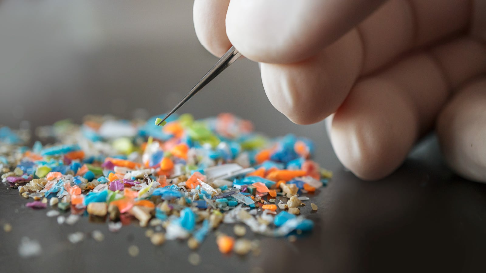  Gloved hand using tweezers to pick up a tiny green fragment from a pile of colorful microplastics on a dark surface.