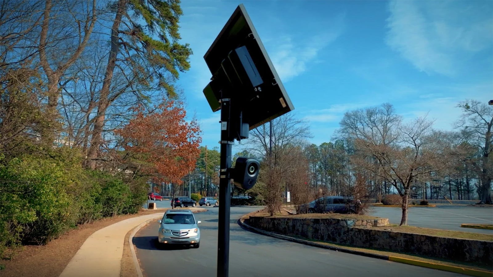 Flock Safety license plate reader camera mounted on a pole beneath a solar panel beside a road, with a car approaching.