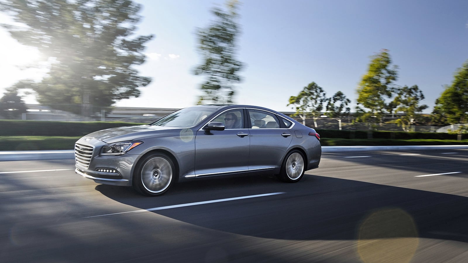 Silver 2016 Hyundai Genesis sedan driving along a tree-lined road, viewed from the front driver’s side.