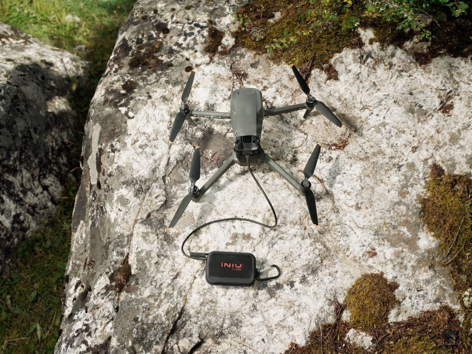 A black INIU Outdoor Rugged power bank with a carabiner clip sits on a lichen-covered boulder, connected by a cable to a gray folding drone with its propellers extended.