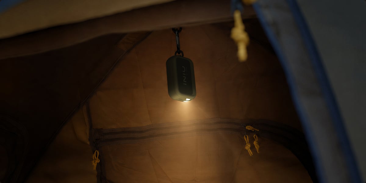 An INIU Outdoor Rugged power bank hangs from the ceiling of a tent by its carabiner, with its built-in light switched on and casting a warm glow downward.