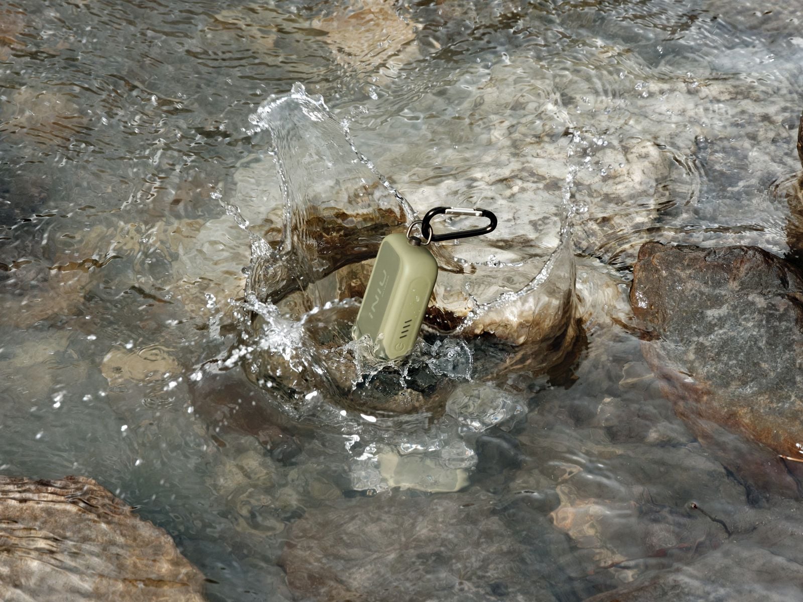 An olive green INIU Outdoor Rugged power bank with a carabiner clip splashes into shallow, rocky stream water, with droplets flying around it.