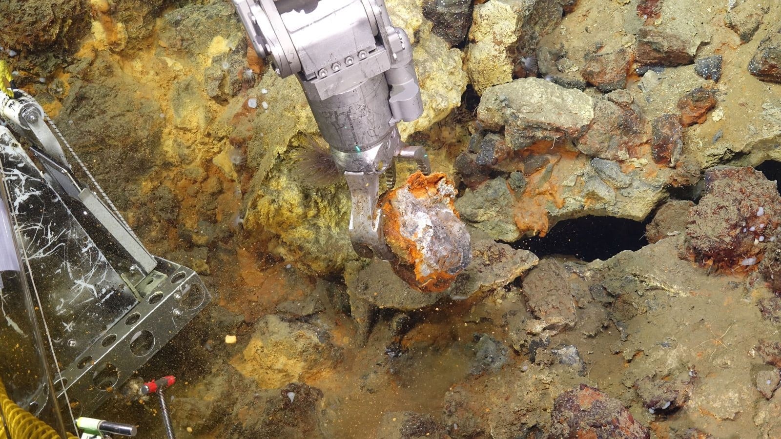 A robotic arm on a remotely operated vehicle grips a rust-orange rock with a silvery interior on the Pacific seafloor, surrounded by yellow and orange mineral-stained rubble.