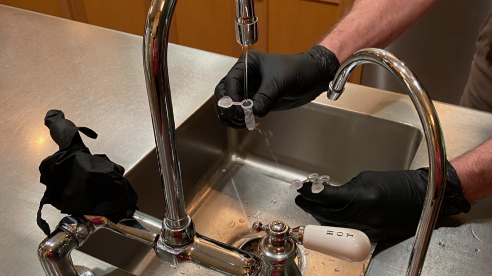 Gloved hands collect tap water from a kitchen faucet into small sample vials for PFAS testing.