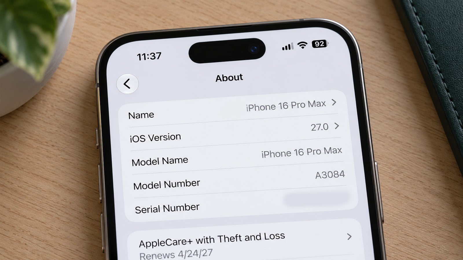 Close-up of an iPhone 16 Pro Max on a desk showing the About screen with iOS 27.0 and device model information.