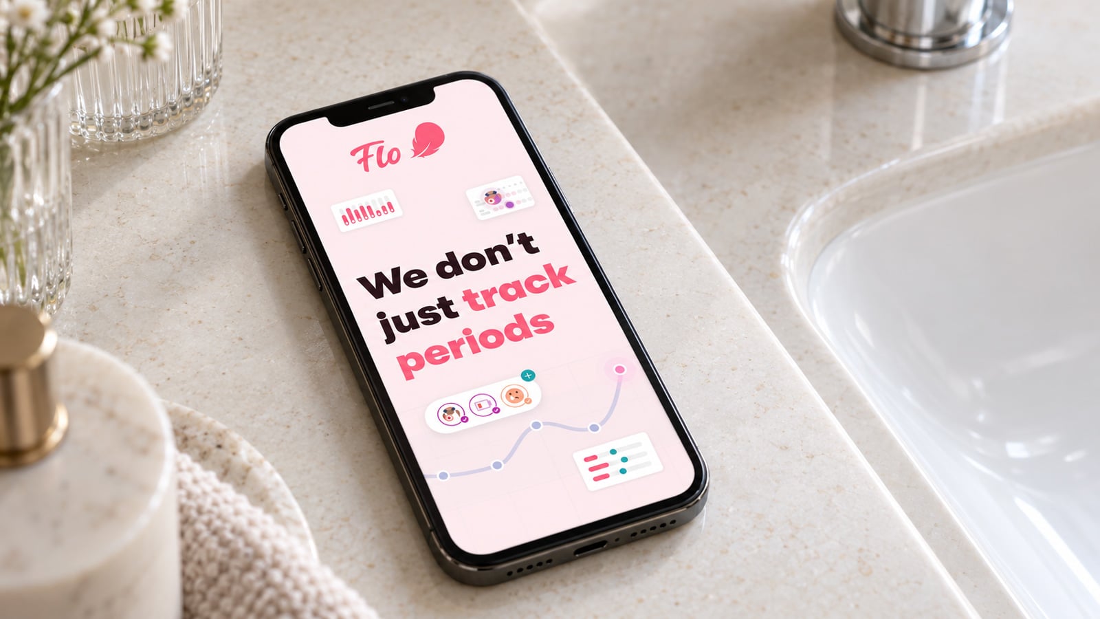 Phone lying flat on a bathroom counter beside a sink, displaying a pink Flo screen with the message 'We don’t just track periods'