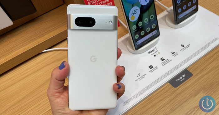 Google Pixel 7 in white held in hand showing the back. You can see additional Pixel 7 models on a white stand on a wood table in the background.