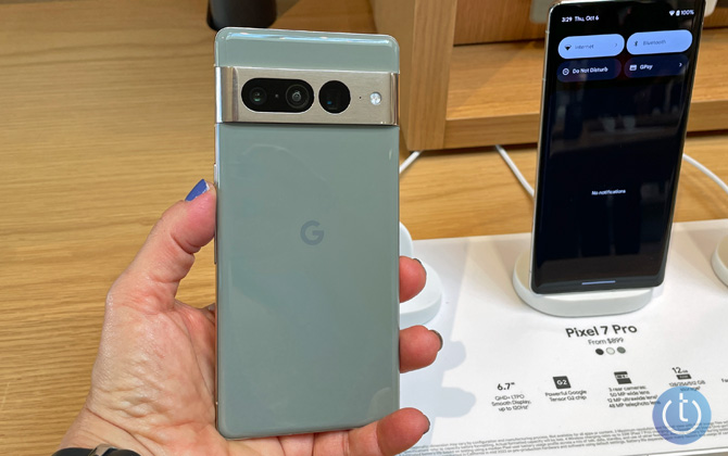 Google Pixel 7 Pro in green held in hand showing the back with two distinct lens clusters. You can see additional Pixel 7 Pro model on a white stand on a wood table in the background.