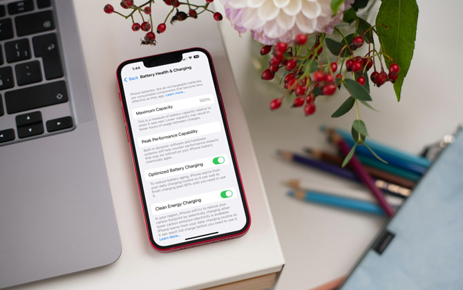 iOS Battery Health & Charging screen shown on iPhone on desk with MacBook and flowers.