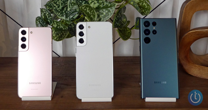 From the left, the Samsung Galaxy S22 (pink), S22+ (white), and S22 Ultra (green) on wooden shelving with plant in the background.